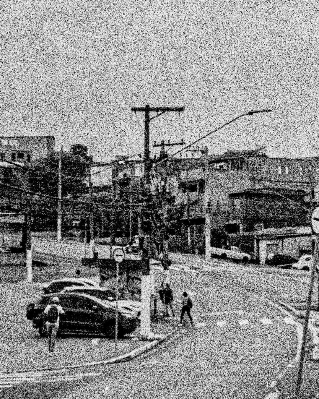 Uma avenida sinuosa com pouco movimento, algumas pessoas e carros aparecem na imagem. A fotografia é PB com bastante ruído e desfoque.