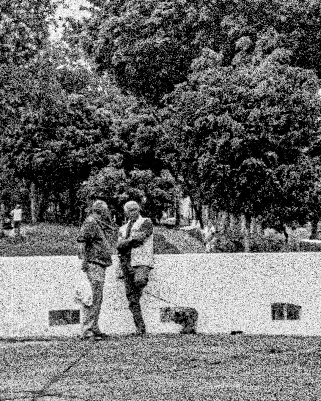 Dois senhores conversando, um deles está com um cachorrinho, ambos estão em frente a um córrego. A fotografia é PB com bastante ruído e desfoque.